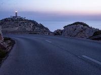 a narrow highway with a lighthouse on a rock cliff above the ocean at dusk overlooking an island