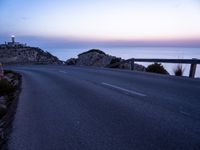 a narrow highway with a lighthouse on a rock cliff above the ocean at dusk overlooking an island