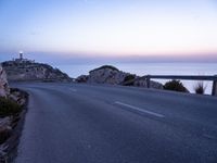 a narrow highway with a lighthouse on a rock cliff above the ocean at dusk overlooking an island