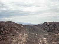 a road covered in dirt on a cloudy day with mountains in the distance on the other side