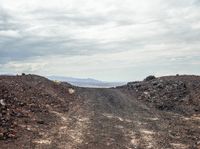 a road covered in dirt on a cloudy day with mountains in the distance on the other side