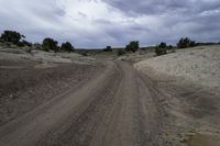 a dirt road through a barren field under cloudy skies with dark clouds in the sky