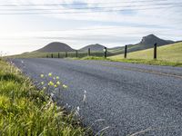 an asphalt road and a fence with some weeds in it, near a grassy hillside