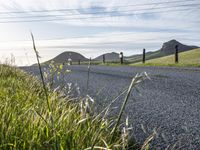 an asphalt road and a fence with some weeds in it, near a grassy hillside