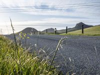 an asphalt road and a fence with some weeds in it, near a grassy hillside