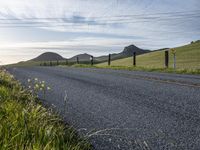 an asphalt road and a fence with some weeds in it, near a grassy hillside