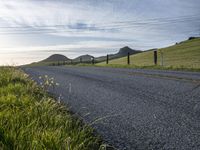 an asphalt road and a fence with some weeds in it, near a grassy hillside