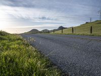 an asphalt road and a fence with some weeds in it, near a grassy hillside