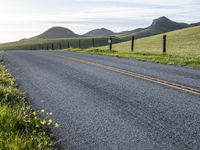 an asphalt road and a fence with some weeds in it, near a grassy hillside