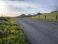 an asphalt road and a fence with some weeds in it, near a grassy hillside