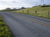 an asphalt road and a fence with some weeds in it, near a grassy hillside