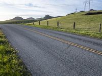 an asphalt road and a fence with some weeds in it, near a grassy hillside