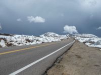 snow covers the roadway and snowy mountains on a sunny day, with a yellow warning sign in front of it