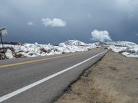 snow covers the roadway and snowy mountains on a sunny day, with a yellow warning sign in front of it