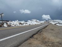 snow covers the roadway and snowy mountains on a sunny day, with a yellow warning sign in front of it