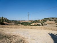 a small hill with trees and grass near a dirt field with an electric pole in the distance