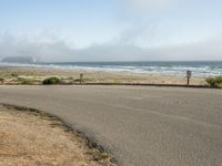 the road leads to an empty beach where two people are sitting and a person is standing near the water