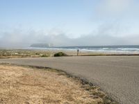 the road leads to an empty beach where two people are sitting and a person is standing near the water