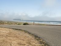 the road leads to an empty beach where two people are sitting and a person is standing near the water