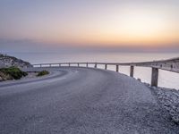 a person standing on the side of a winding road over looking the ocean at sunset