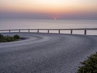 a person standing on the side of a winding road over looking the ocean at sunset