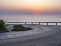 a person standing on the side of a winding road over looking the ocean at sunset