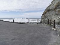 a road going past a rock beach with waves coming in to the shore area and a person walking along the side