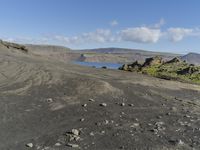 a rock covered beach surrounded by hills under a blue sky on a windy day's day