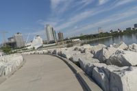 there is a rock wall lined walkway by the water's edge with a city skyline in the distance