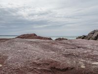 a man stands at the edge of a rocky beach while standing on red sand and looking out into the ocean