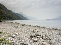 a rocky beach is covered in debris and grass by water and mountains under clouds a sign that reads a no swimming zone