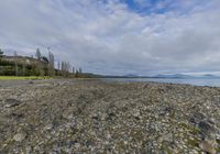 an empty rocky beach near a large body of water and hills in the distance with a few clouds above