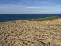 a person standing on a rocky beach near the ocean next to a sandy hill with a dog sitting in it