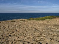 a person standing on a rocky beach near the ocean next to a sandy hill with a dog sitting in it