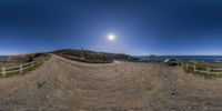 a dirt bike ramp is on the rocky beach beside the ocean with a beautiful full moon in the blue sky