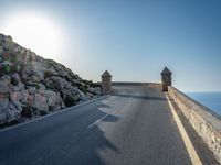 a curve of road leading towards a rocky cliff, with the sky shining through a clear blue sky