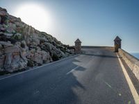 a curve of road leading towards a rocky cliff, with the sky shining through a clear blue sky