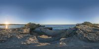 the sun rises over rocks at the beach with the ocean behind them as a fisheye lens