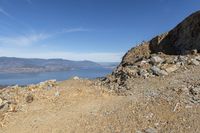 a rocky hillside with a lake and mountains behind it, with a man looking out at the water from high up in the sky