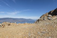 a rocky hillside with a lake and mountains behind it, with a man looking out at the water from high up in the sky