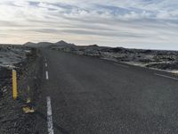 the asphalt road has three yellow posts beside it and hills in the distance behind it