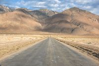 a large open country road in the desert with mountains behind it and a cloud filled sky above