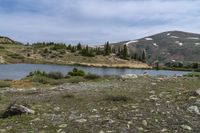 the grassy hillside near a river is rocky and has a lake on it with snowcapped mountains in the distance