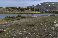 the grassy hillside near a river is rocky and has a lake on it with snowcapped mountains in the distance