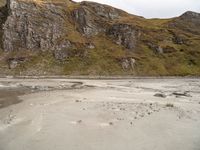 a horse standing in the dirt at the bottom of a mountain near water and rocks