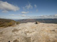 man with a white board standing on a large rock looking at the scenery of a valley