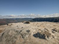 the sky is clear and blue with white clouds above, a lone rock sits on the top of a mountain