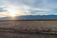 an open plain with the sun peeking over the mountains in the distance and the sun setting on the horizon