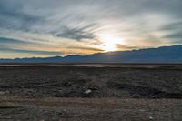 an open plain with the sun peeking over the mountains in the distance and the sun setting on the horizon