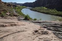 a river flows in the distance next to a cross on a rocky trail as two people walk